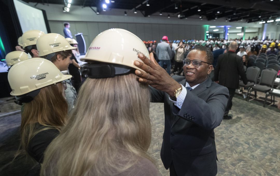 Man places a hard hat on a female student's head at the front of a large room.
